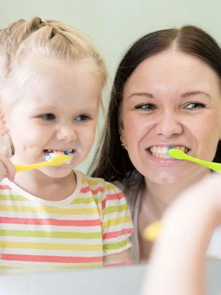 Adult and young child brushing teeth together with yellow toothbrushes, smiling.