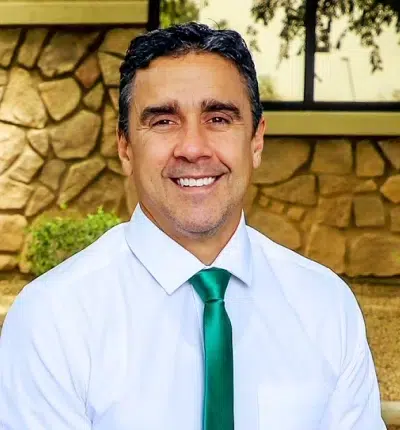Professional headshot of smiling man in white shirt and green tie against stone wall background.